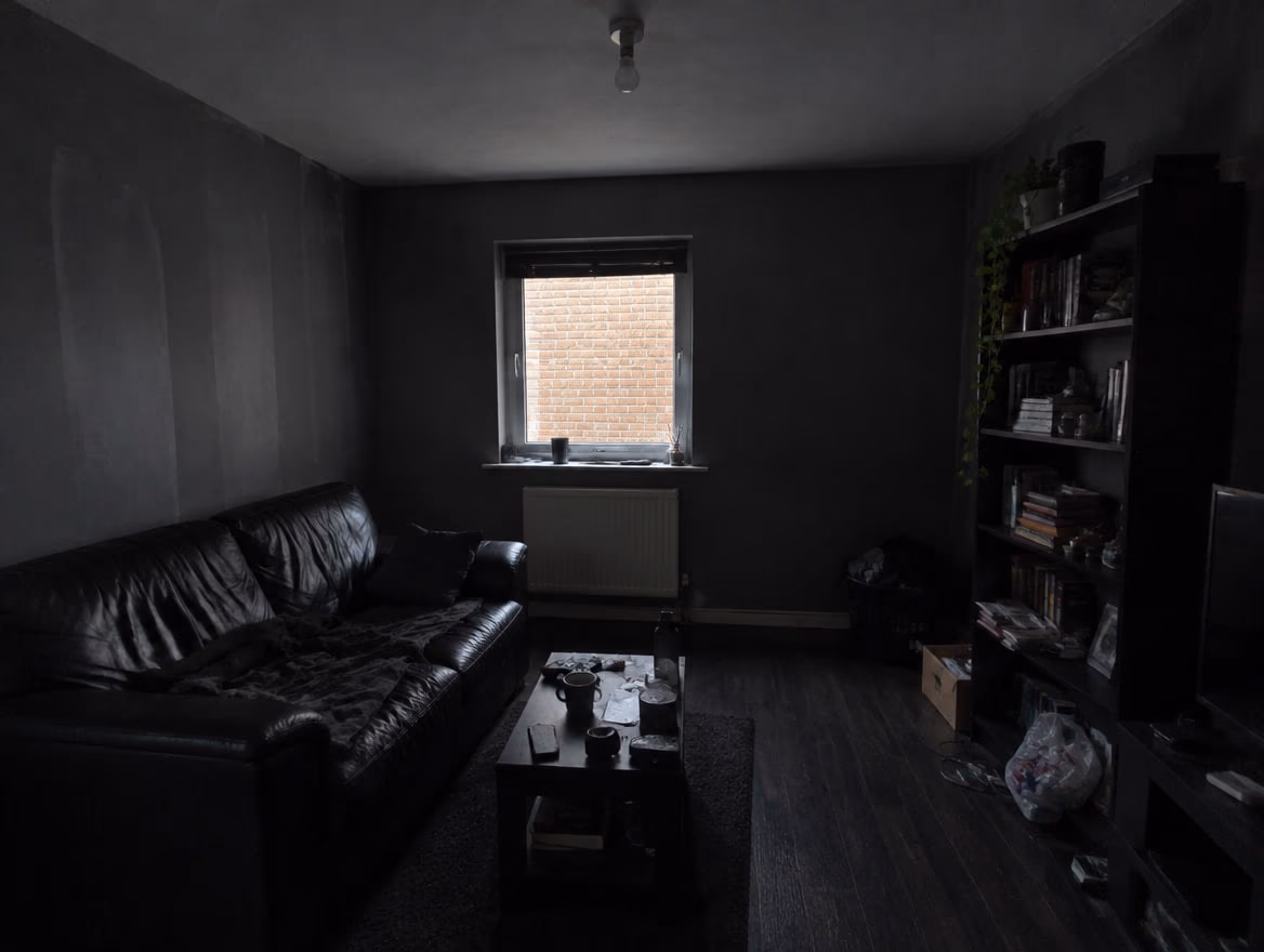 A dim living room with one small window facing a wall, dark gray walls and a black leather sofa