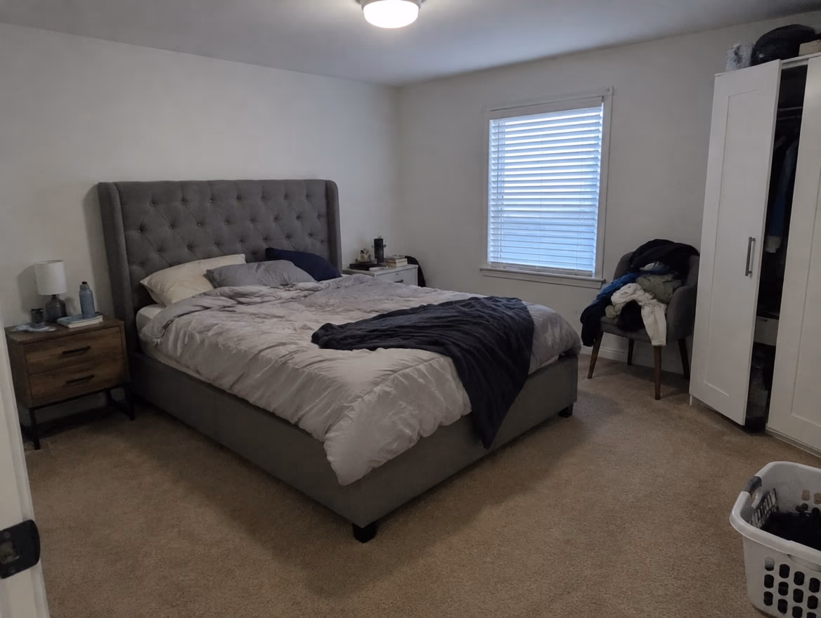 An ordinary bedroom with a tall gray tufted bed, mismatched nightstands and clothes on a chair