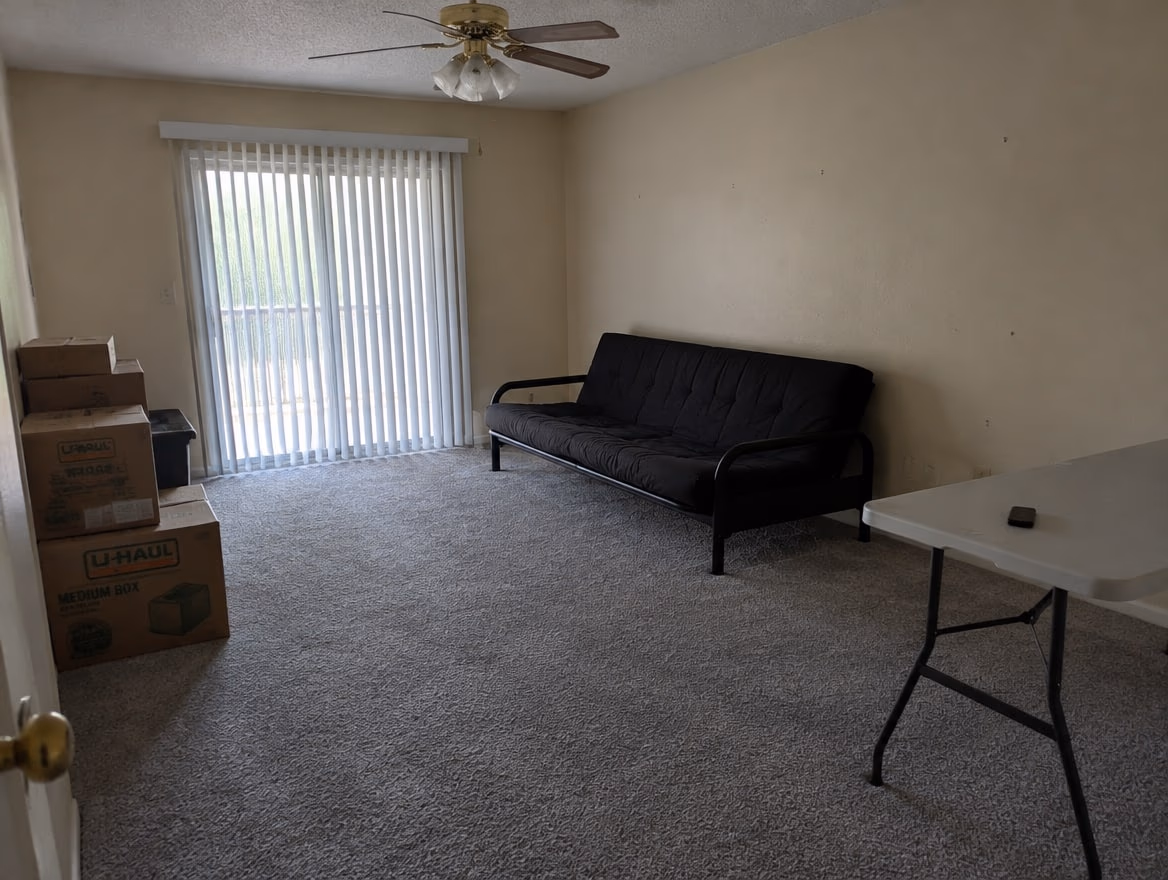 A bare rental living room with beige walls, gray carpet, vertical blinds and a futon