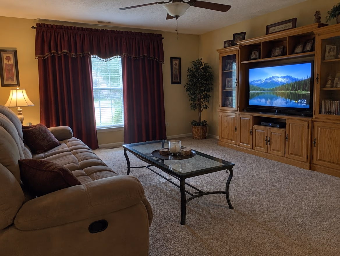 An ordinary living room with a tan reclining sofa, burgundy drapes and an oak entertainment center