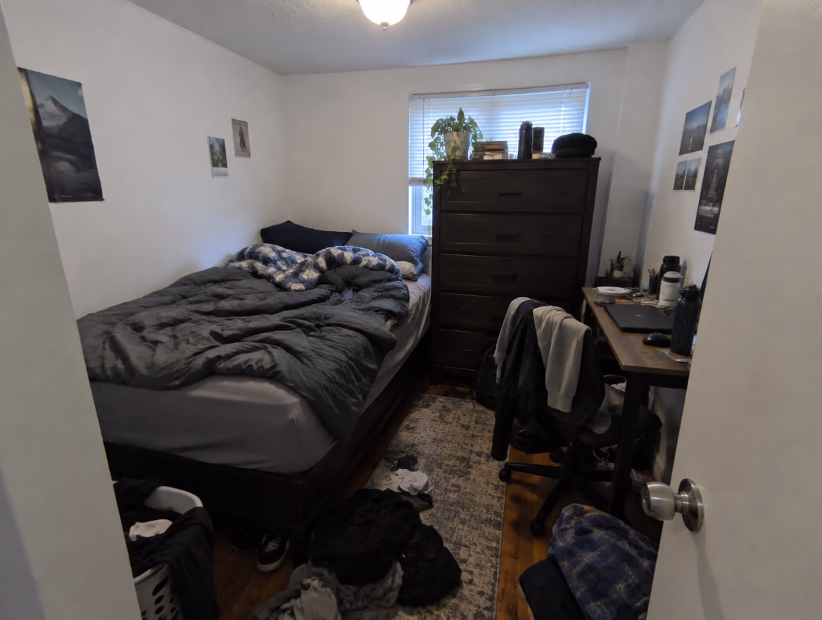 A tiny cramped bedroom with the bed in the corner, a dresser blocking the window and clothes on the floor